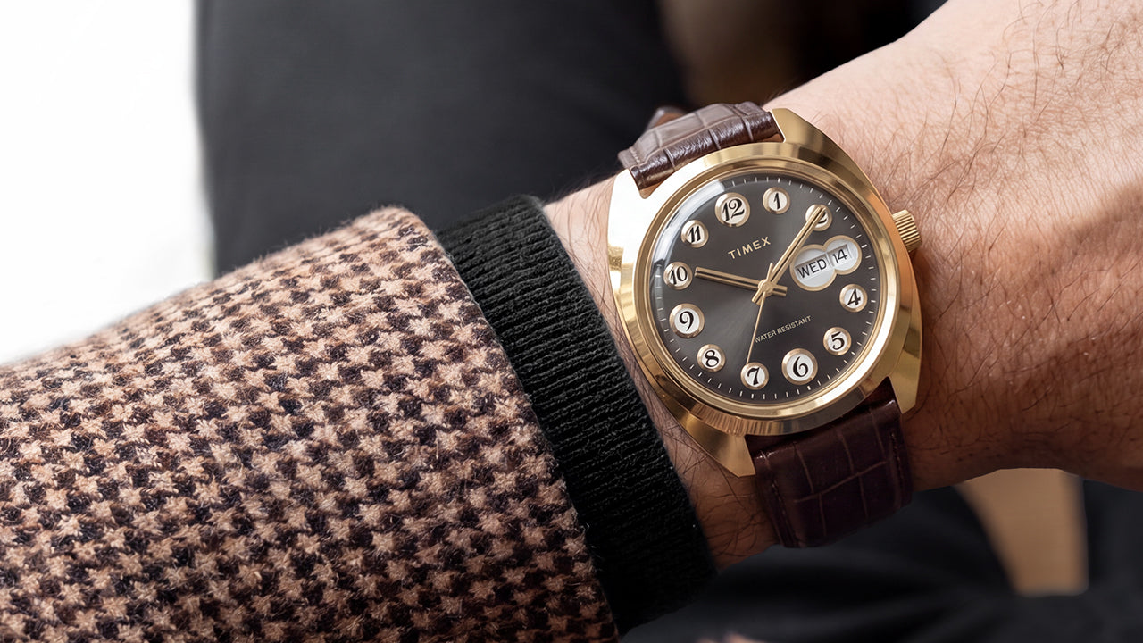 Timex 1976 Rotary Phone Dial Reissue: tárcsás telefon a csuklón Timex® 1976 Rotary Phone Dial Reissue with a gold-tone case, black dial, and brown leather strap worn on the wrist with a patterned blazer.