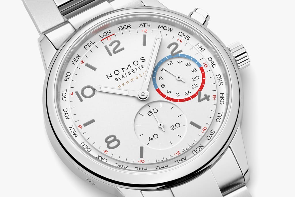 Close-up of a NOMOS worldtimer watch dial with city ring, small seconds subdial, and silver case on a metal bracelet.