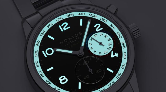 Nomos Club Sport Neomatik Worldtimer Dark: világidő fekete számlapon, teljes fényerővel Close-up of a NOMOS watch face with turquoise bezel ring showing city codes, black dial, cyan luminescent numerals and hands, metal bracelet.
