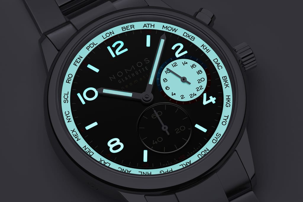 Close-up of a NOMOS watch face with turquoise bezel ring showing city codes, black dial, cyan luminescent numerals and hands, metal bracelet.