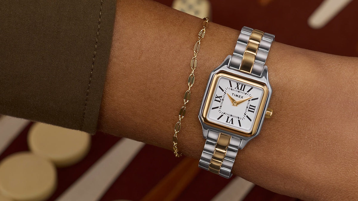 A kis órák visszatérnek – de nem mindegy, hogyan viseled őket Timex Legacy Addison two-tone square watch with white Roman-numeral dial, worn on the wrist beside a delicate gold chain bracelet.