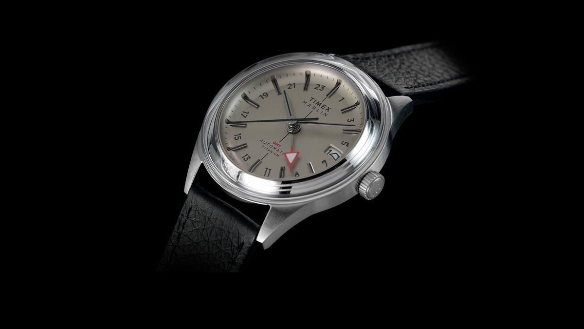 Timex Marlin Jet Titanium Automatic GMT: retrofuturisztikus tok, titánból Angled view of the Timex Marlin® Jet Titanium Automatic GMT featuring a brushed titanium case, cream dial, black leather strap, and red GMT hand.