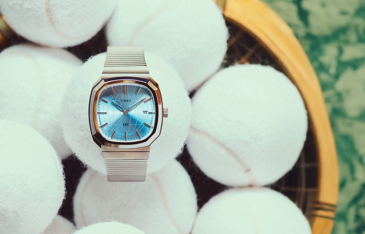 END. x Timex 1981 Reissue Inspired watch with a stainless-steel case, blue globe-pattern dial, and expansion bracelet, displayed among tennis balls.