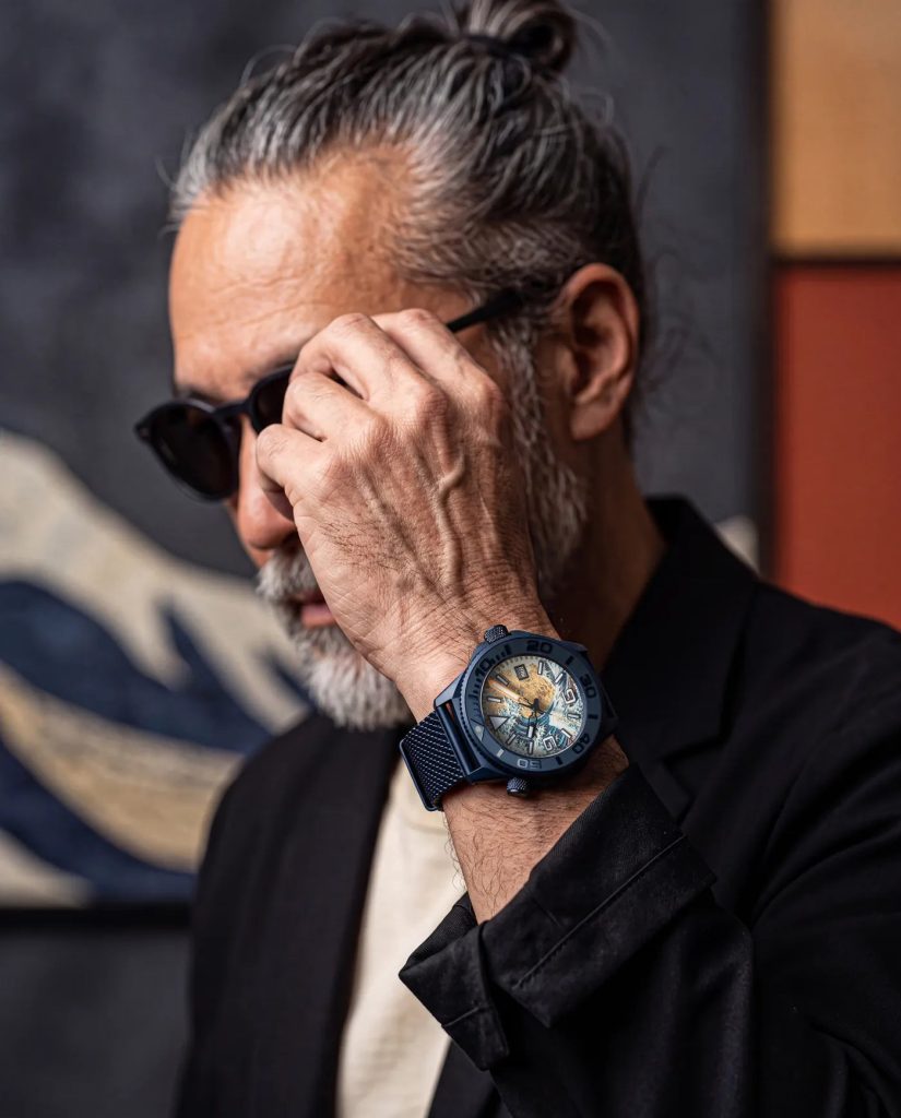 UNDONE Aquadeep The Great Wave - Visszatért és most aranyfüstre épül Hokuszai nagy hulláma Az óra fekete zakóhoz és napszemüveghez viselve, hullámos falikárpit előtt