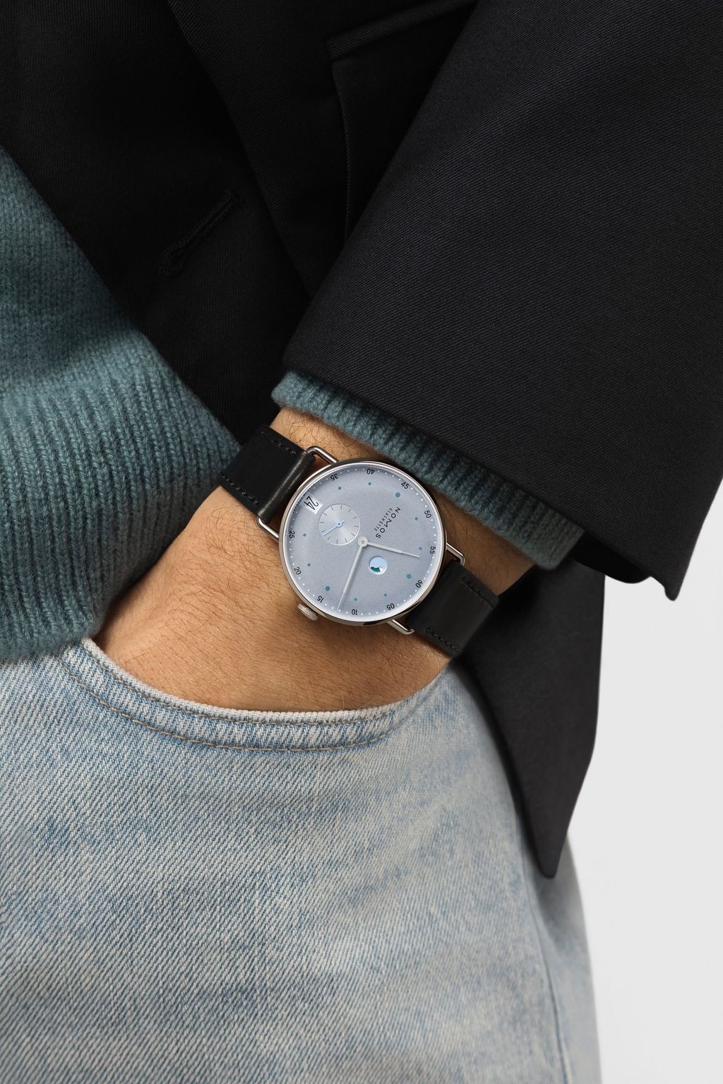 Jégkék Metro új kézi felhúzású kaliberrel: bemutatkozik a Nomos DUW 4701 Wrist shot of a NOMOS watch with ice blue dial and black strap, worn with jeans and a dark jacket.