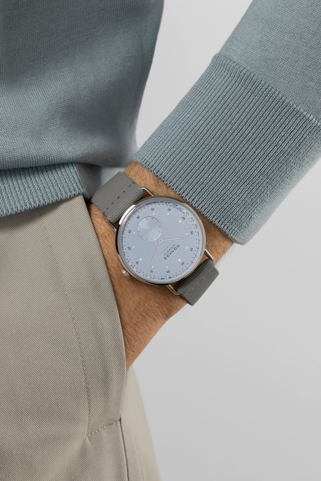 Jégkék Metro új kézi felhúzású kaliberrel: bemutatkozik a Nomos DUW 4701 Wrist shot of a NOMOS watch with ice blue dial and gray fabric strap on a person wearing a light blue sweater and beige trousers.